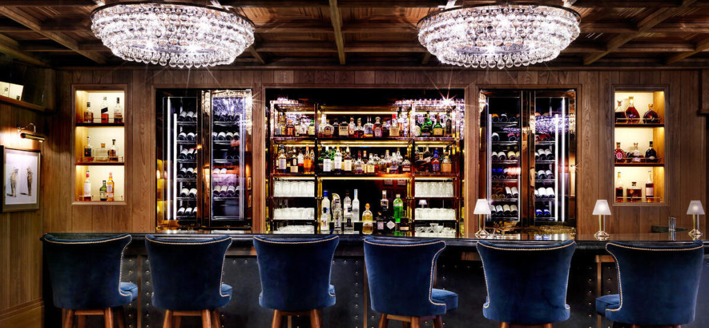 A luxurious bar interior with plush blue barstools faces a well-stocked, illuminated wooden bar. Two large, elegant crystal chandeliers hang from the ceiling, complemented by brass bar shelving that displays various bottles and glasses. Cozy and inviting ambiance.