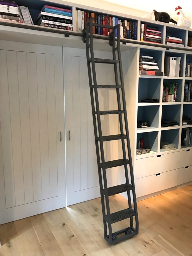 A steel rolling ladder leaning against a bookshelf filled with various books. The bookshelf is adjacent to two closed doors and a section of open shelving. A wooden floor is visible beneath the setup.