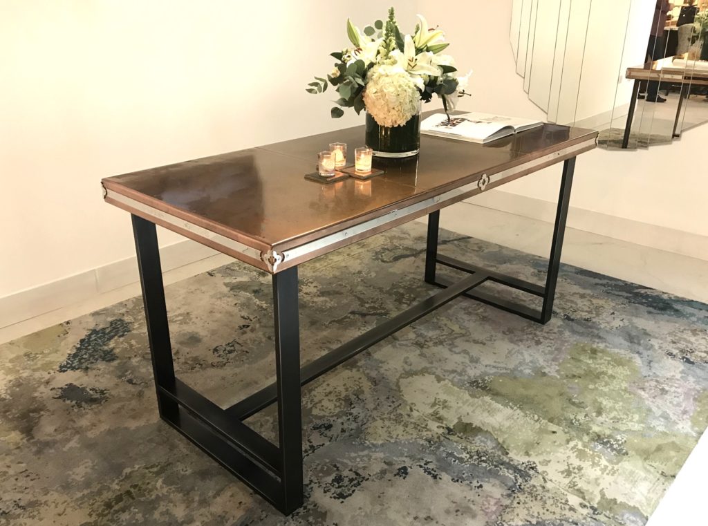 A sleek, modern copper table is set on a patterned rug. It features two lit candles and a floral arrangement in a vase, with a book open beside them. A mirror reflects the scene in the background.