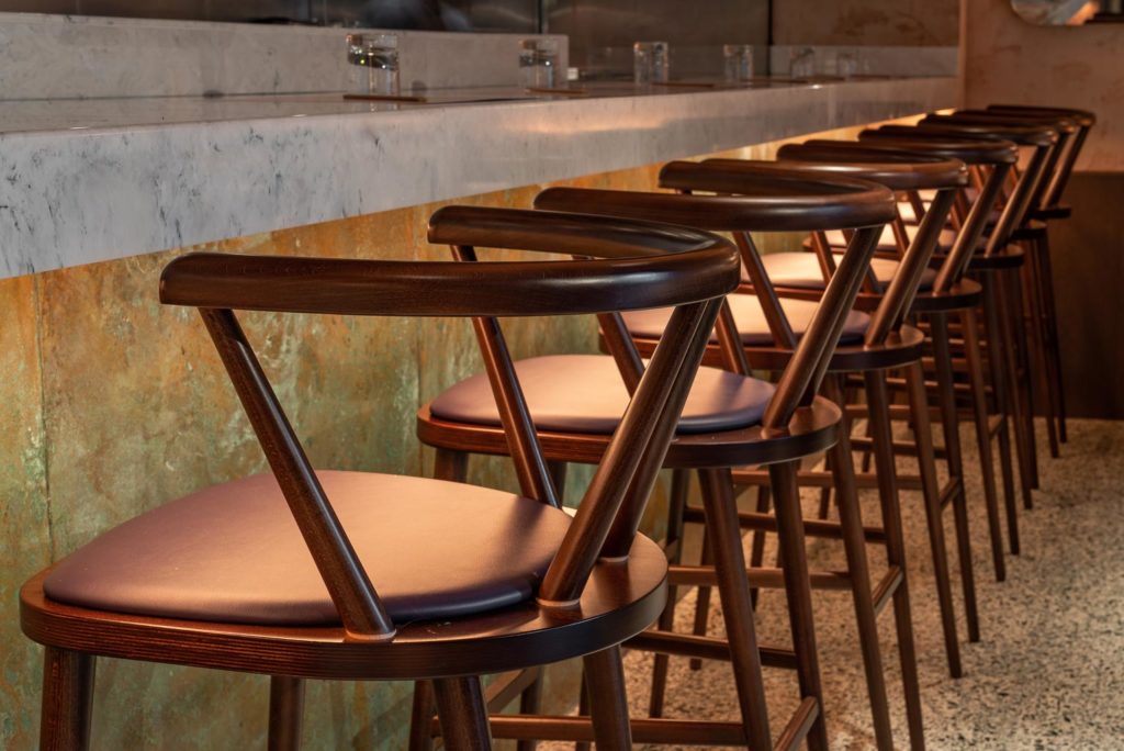 A row of stylish wooden barstools with curved backs and brown leather seats is neatly arranged at a marble-topped bar counter. Patinated copper panels add an artistic touch, as warm lighting enhances the cozy atmosphere, reflecting off the polished surfaces.