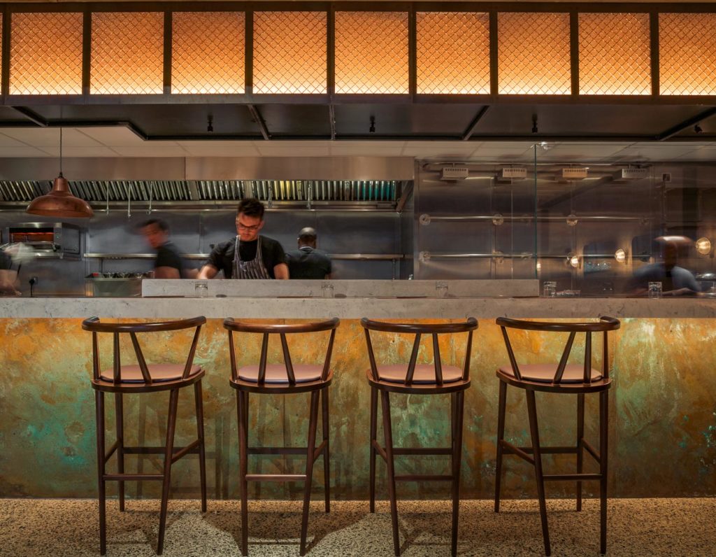 A dimly lit restaurant kitchen features a counter with four high wooden chairs, while patinated copper panels add a touch of elegance. A chef in an apron expertly works behind the counter, as blurred staff move energetically in the background, enhancing the warm, industrial ambiance.