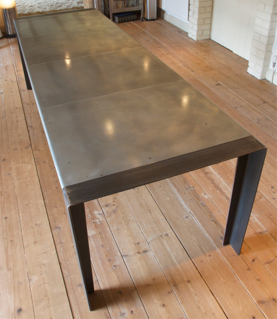 A long, modern zinc top dining table with a smooth metallic surface and sleek, dark legs stands on a wooden floor in a room with white brick walls.