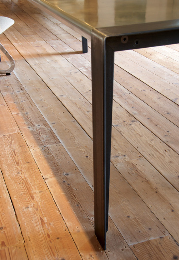 Close-up of a bespoke metal table leg with a zinc tabletop on a wooden floor. The table leg boasts a sleek, modern design. Part of a chair and the floor’s natural grain are visible as soft shadows dance across the floorboards.