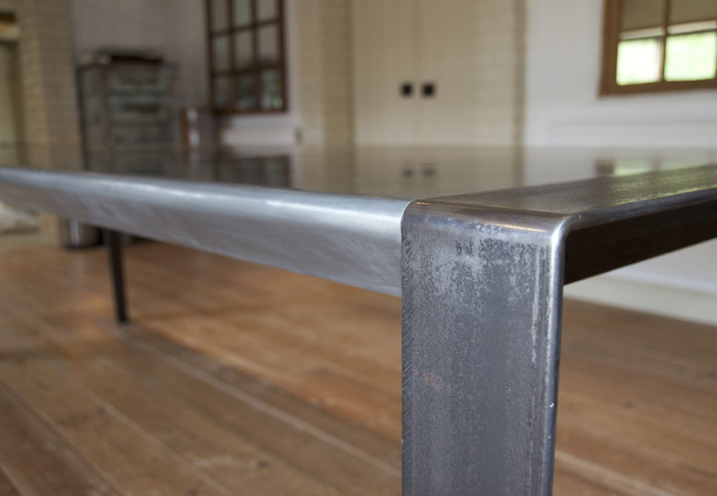 Close-up of a minimalist industrial-style metal large dining table, featuring a smooth surface and solid legs on a wooden floor. The background showcases large windows and blurred interior features, conveying a modern, spacious room.