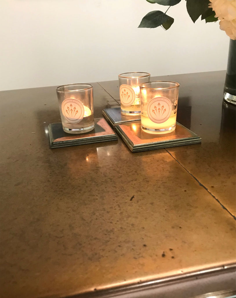 Three lit candles in glass holders sit on coasters on an antique copper table. Each candle holder boasts a floral design, and part of a floral arrangement is visible in the upper right corner. The tables warm, reflective surface adds a touch of elegance to the scene.