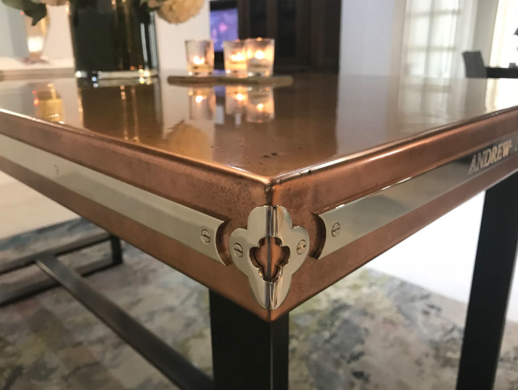 Close-up of a copper wine table corner with a gleaming top and metallic decorative edge. The shiny surface reflects nearby candles and the rooms soft lighting. This elegant table, perfect for any gathering, has sturdy black legs resting on a colorful carpet.