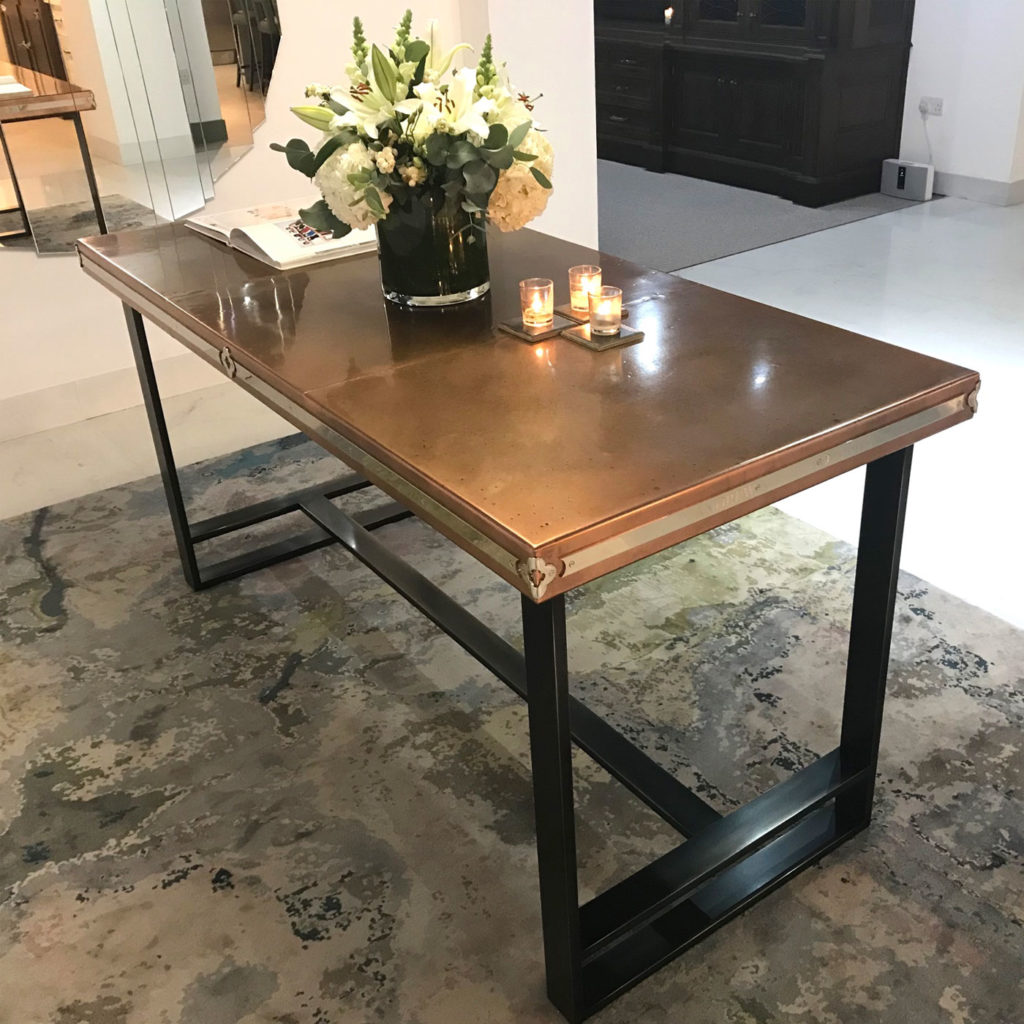 A sleek, bespoke copper table with a metallic top and black frame stands on a patterned rug. A vase of white flowers and two lit candles adorn the table, adding an elegant touch. A book lies open on one side.