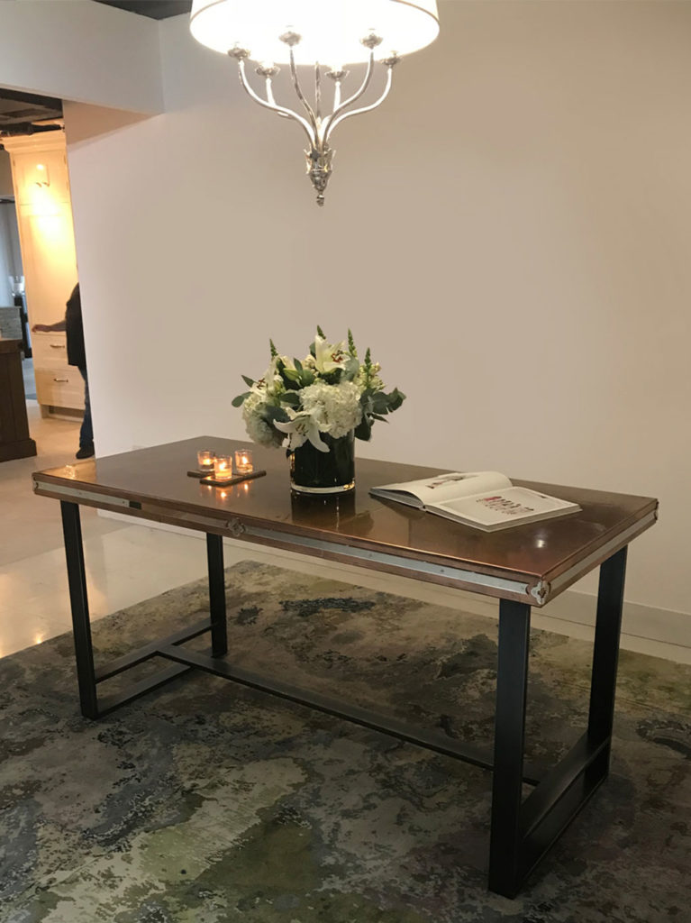 A room with a copper console table showcasing a floral centerpiece, two lit candles, and an open book. A chandelier hangs above, casting light on a patterned rug below.