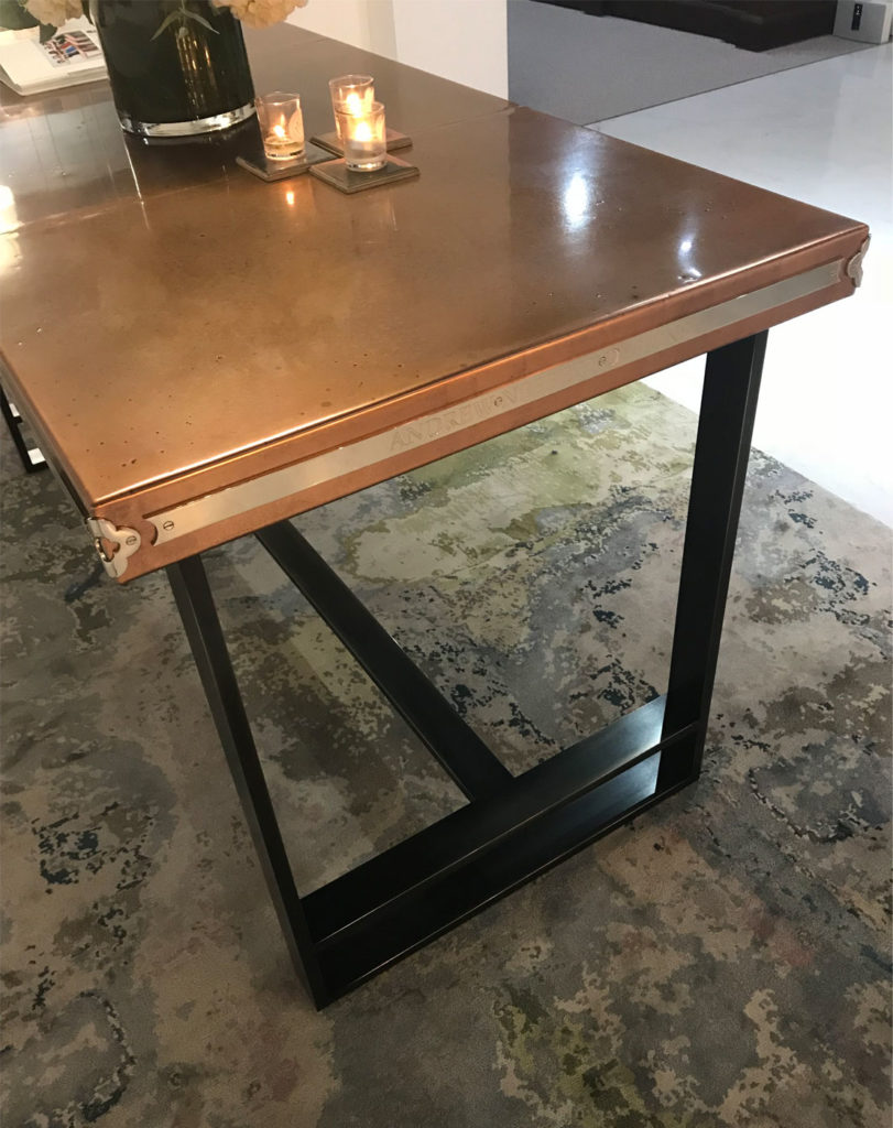 A bespoke modern table with a polished copper top and black metal legs stands elegantly. Two small lit candles sit at one end near a dark green vase, resting on the patterned marble floor, embodying an industrial charm.