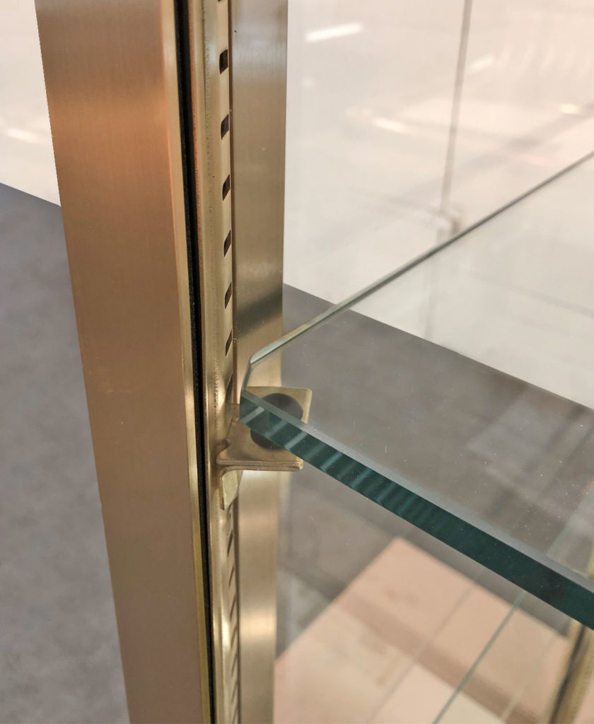 Close-up of a bespoke brass and glass mirror cabinet corner with metallic support and adjustable slots. The transparent shelves, crafted from glass, boast a reflective surface. In the blurred background, a light-colored floor subtly complements the elegant design.