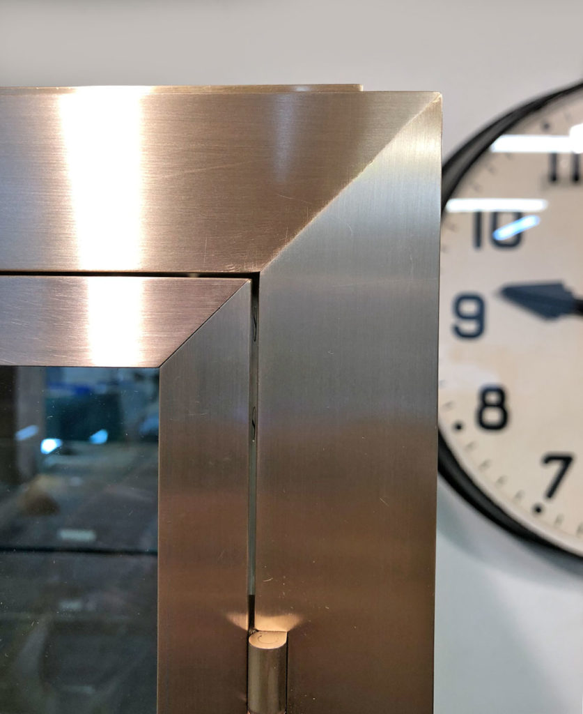 Close-up of a bespoke metal-framed corner of a glass display case, with a wall clock showing 11:58 in the background. The brushed brass surface reflects light, echoing the craftsmanship and design typical of an exquisite glass mirror cabinet.