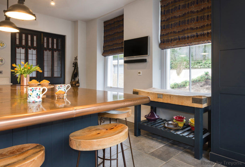 A modern kitchen boasts a large cooper island with colourful mugs and flowers. Wooden stools surround the space, enhanced by a sleek copper kitchen worktop. Two windows with patterned blinds frame the room, while a mounted TV and a decorative table with plants and bowls add charm below.