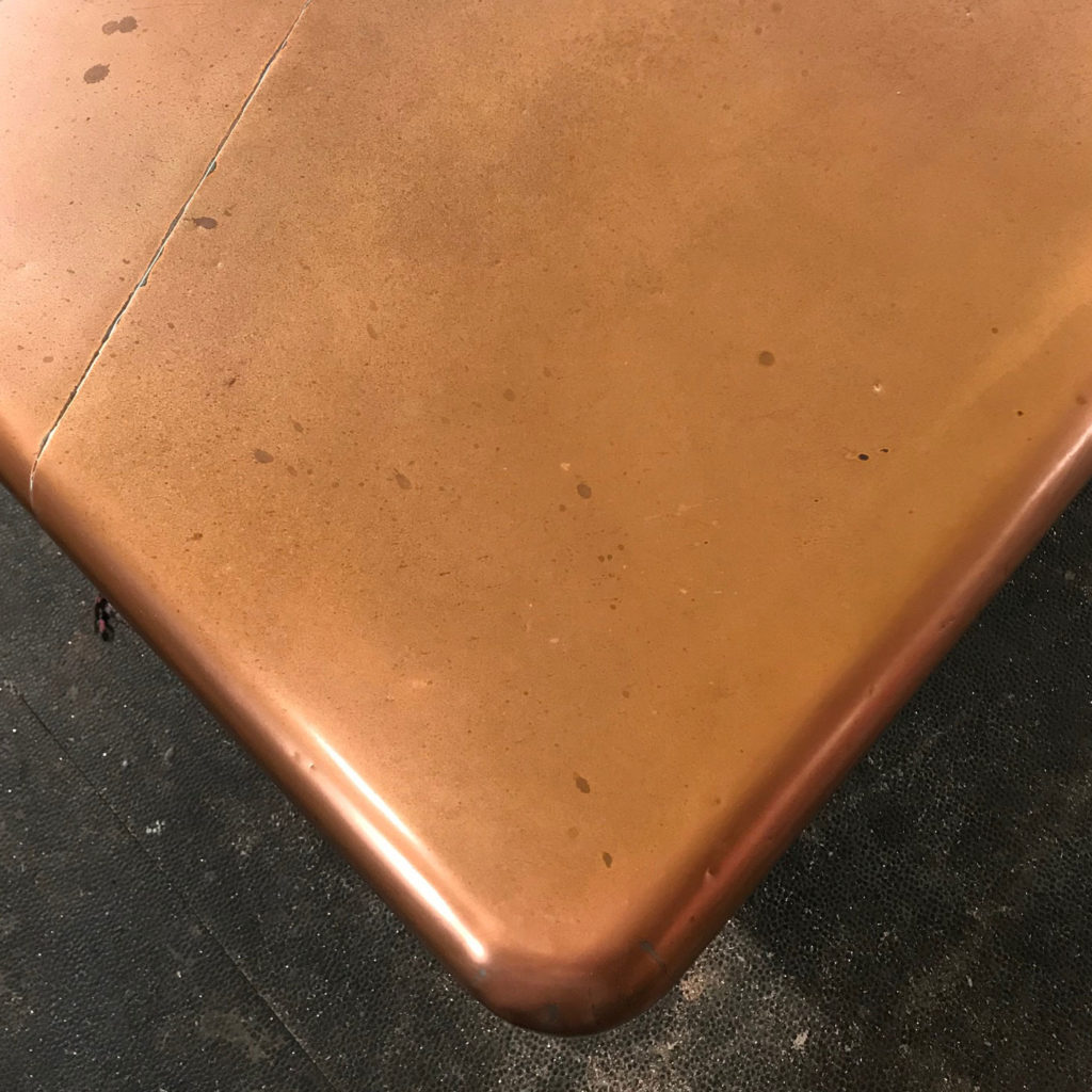 A close-up of a copper kitchen island corner with a rounded edge, showcasing a few small stains on its surface. The island is set against a dark, textured background.