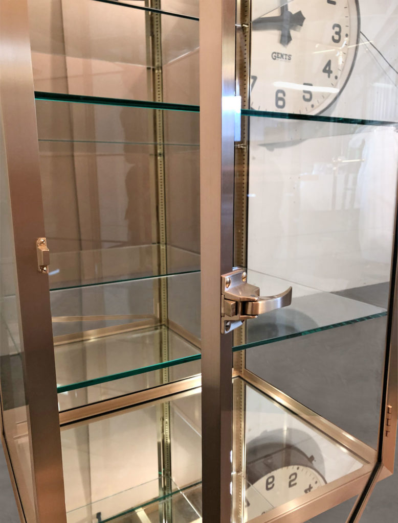 A bespoke glass display cabinet with a metallic frame and four shelves stands open. Inside, a large vintage-style clock boasts black numbers and hands on a white face. The brass details of the cabinet reflect the surrounding light softly like a mirror.