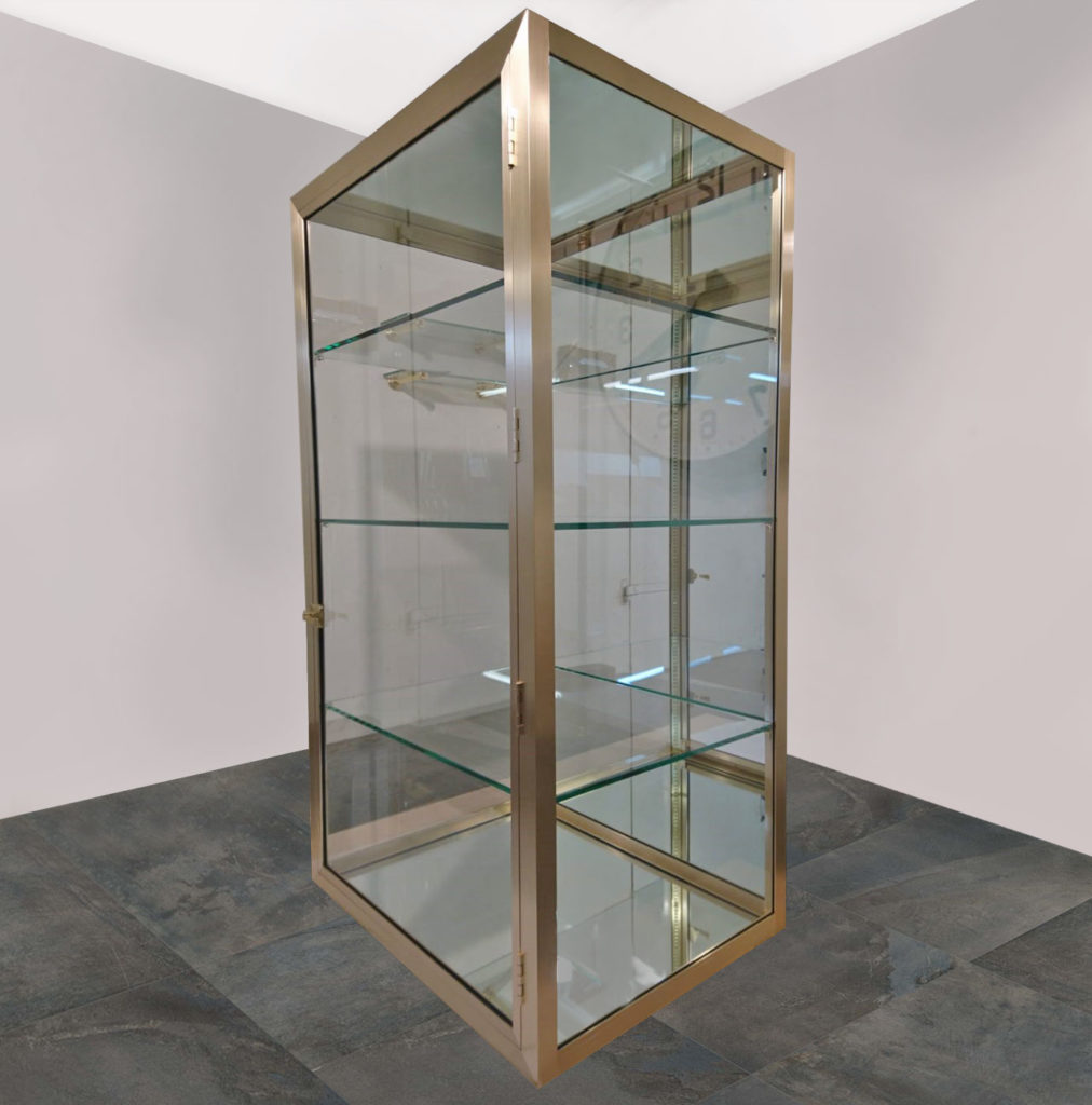 A tall, rectangular bespoke brass and glass mirror cabinet with a metallic frame stands on a stone tile floor. The case features multiple glass shelves and transparent sides, providing visibility from all angles. A simple, light gray wall serves as the perfect backdrop.