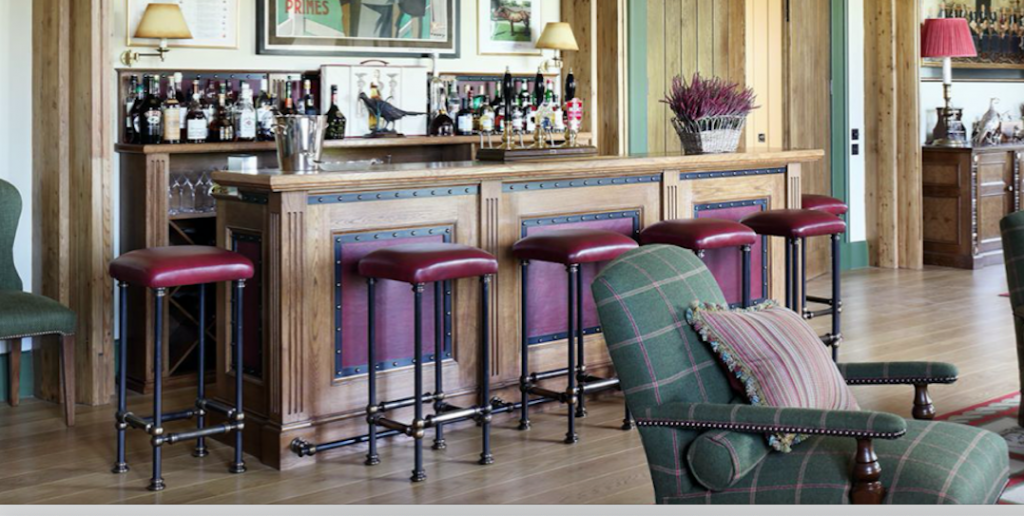 A cozy home bar with wooden paneling and a selection of bottles displayed on shelves. Custom, red-cushioned bar stools line the counter, and a green plaid armchair is in the foreground. Warm lighting and framed pictures adorn the walls.