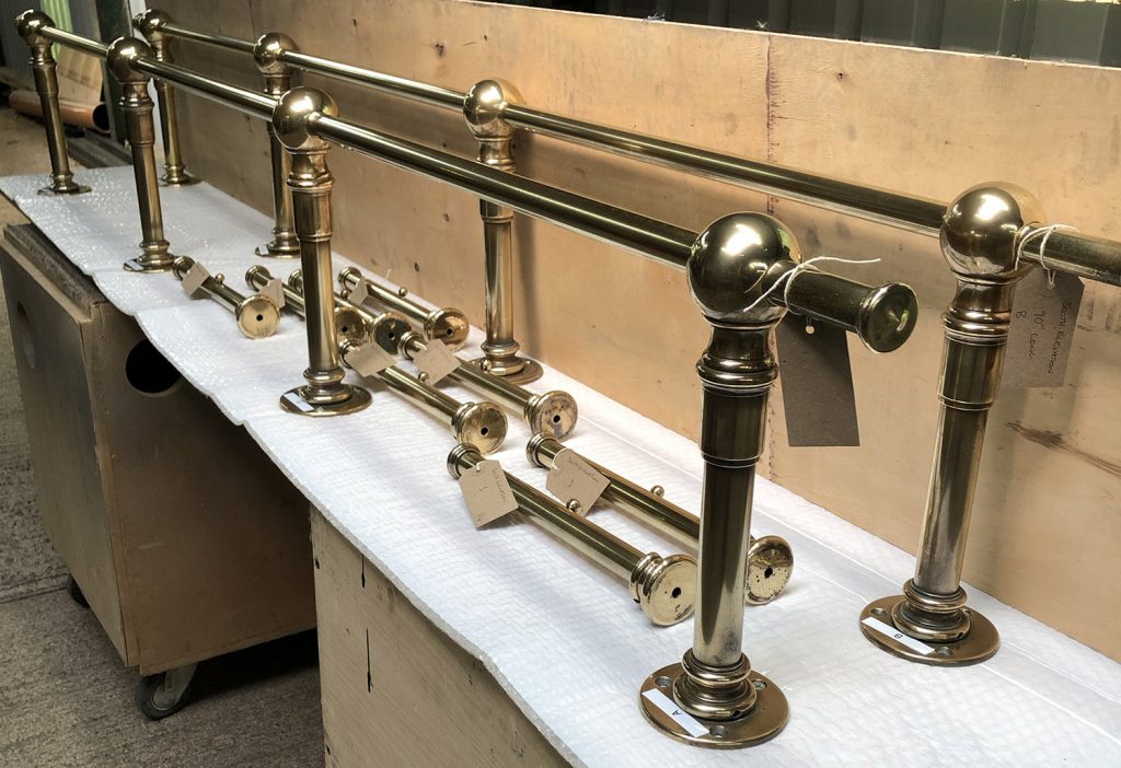 A row of polished brass safety bar posts and components, reminiscent of bespoke dressing room furniture, are arranged on a padded surface. Each post features a round base and spherical finials, with some lying disassembled. Tags adorn certain pieces against the backdrop of wooden boards.