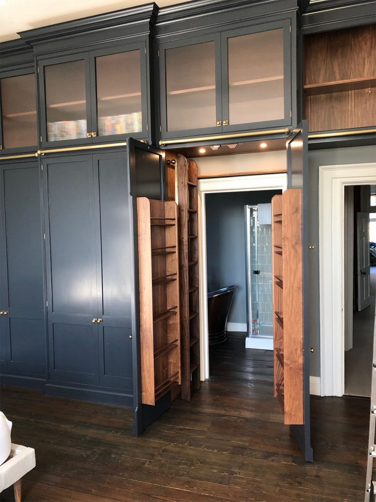 A room features dark blue built-in cabinets with hidden doors that open to reveal a secret passage. The bookshelf-style doors boast wood accents and can be accessed using a collapsible walnut ladder. The wooden flooring enhances the charm as natural light filters in softly.