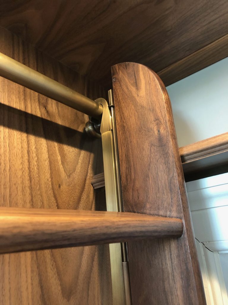 Close-up of a collapsible wooden ladder with brass-colored metal rails, elegantly attached to a walnut-toned wall or shelving unit. The rich, dark grain complements the shiny finish of the metal, offering a stylish touch perfect for any dressing room setting.