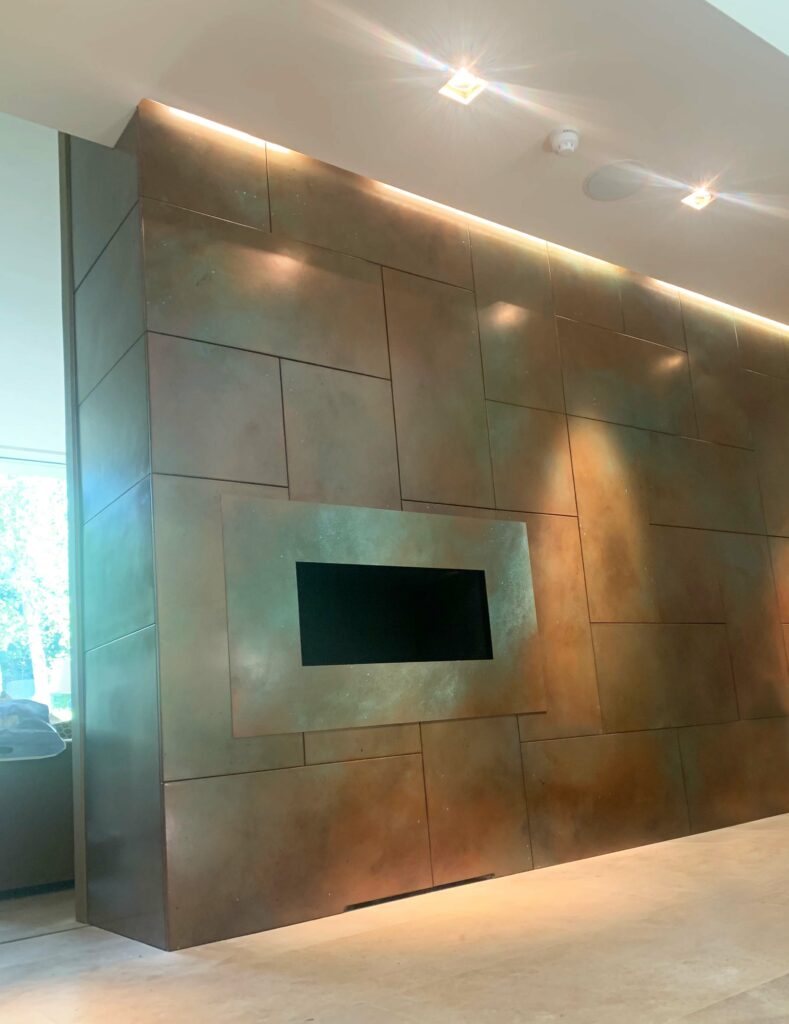 A modern, geometric wall with metal panels and a copper-toned finish features a rectangular inset display. Ambient lighting highlights the metallic textures, creating a sleek, contemporary atmosphere. A large window on the side allows natural light in.