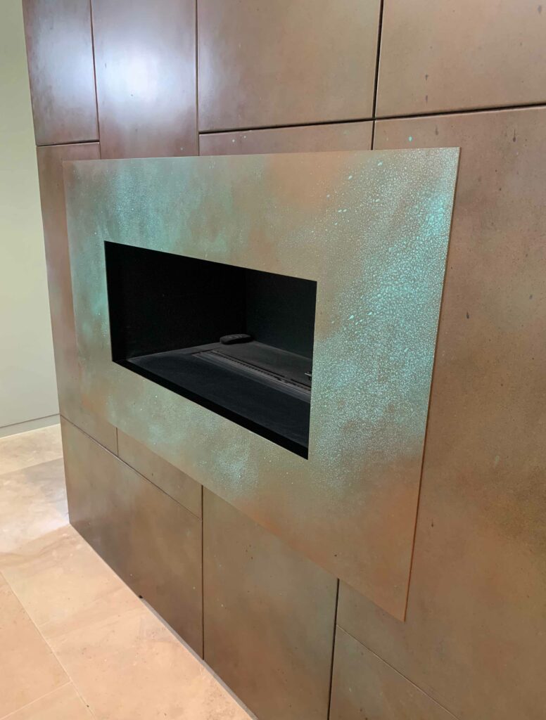 A custom fireplace with a rectangular opening is set into a wall adorned with large, rectangular copper panels. The surrounding area features a slightly green patina, adding texture and contrast to the design. The floor's light-colored tile enhances this modern aesthetic.