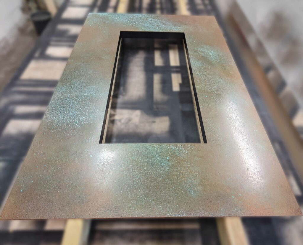 A rectangular copper fire surround displays a textured patinated surface in shades of blue, green, and rust. It rests on a wooden platform with shadows elegantly cast around it.