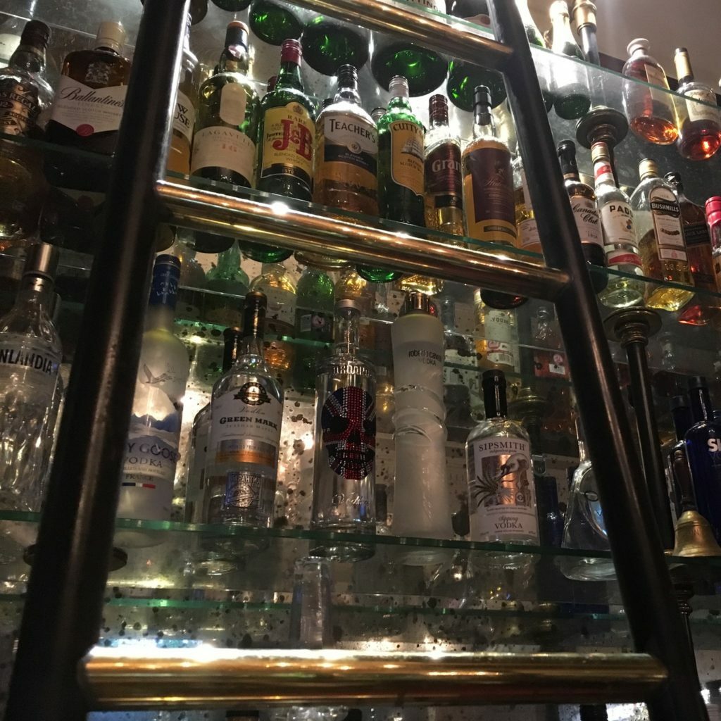 A tall ladder stands before a mirrored bottle display in a bar, stocked with various liquors. The shelves, filled with colorful bottles, reflect the dim lighting, casting a warm glow on the glass and labels, creating an inviting atmosphere.