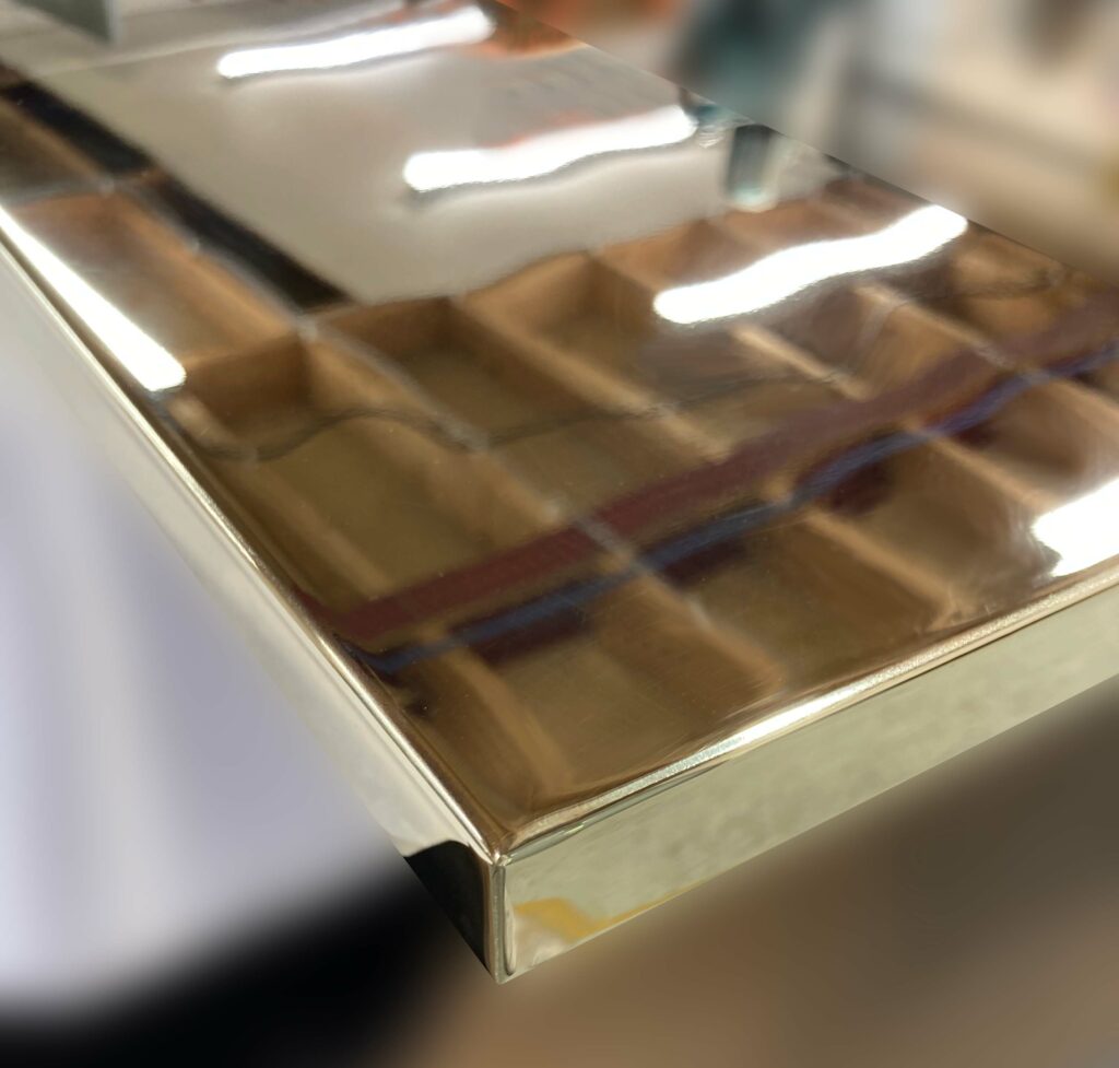 A close-up of a shiny, reflective metal polished brass kitchen cabinet doors with a smooth, mirror-like finish. The background is blurred, highlighting the polished edge of the metal.