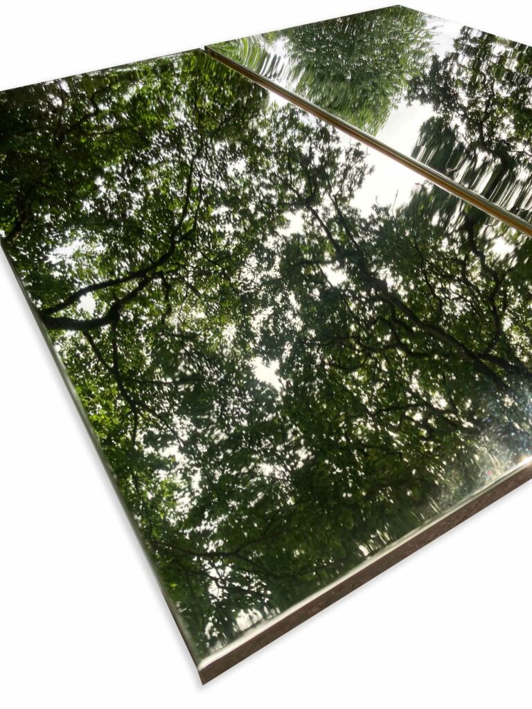 Two rectangular polished kitchen cabinets, framed by custom-made polished brass are placed side by side. They reflect tree branches and green leaves, creating an illusion of looking up into a lush forest canopy.