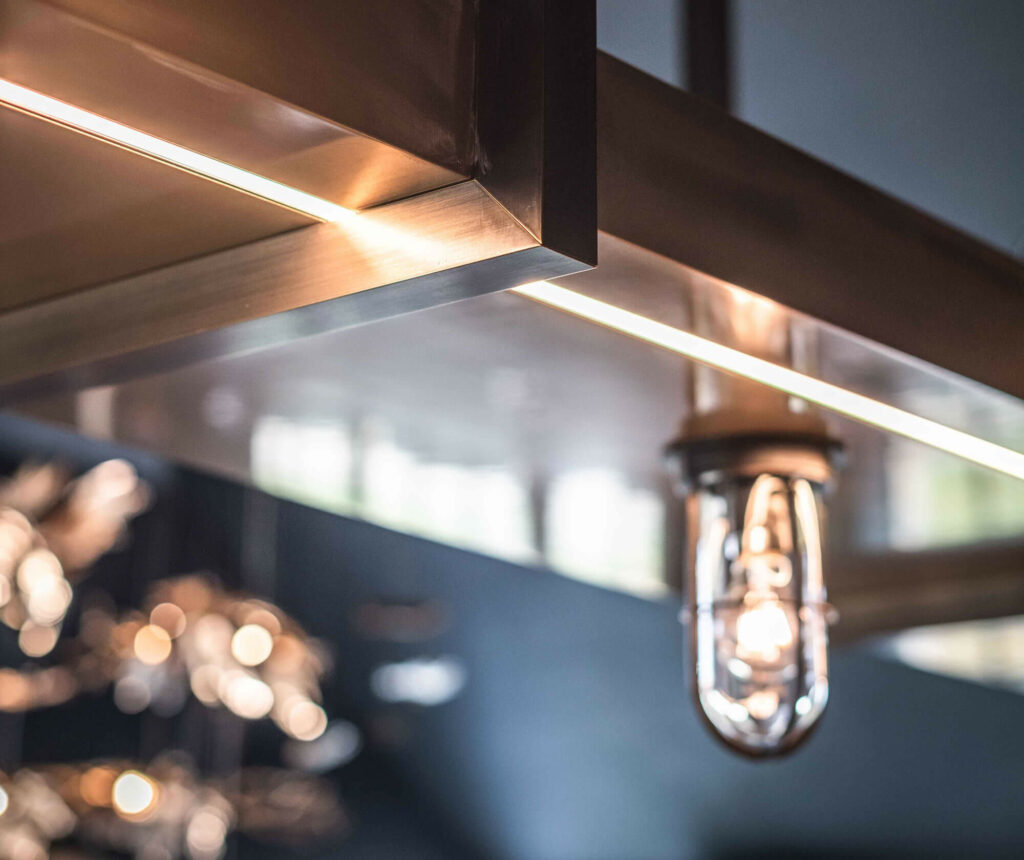 Close-up of a modern metallic light fixture with a glowing bulb, set against a dark blurred background. Soft reflections and highlights are visible on the metal surface.