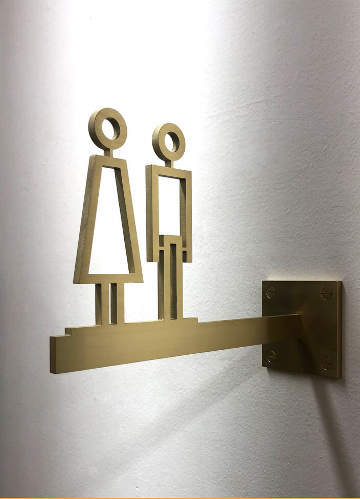 A brass sign with cut out male and female symbols is mounted on a textured wall. The polished surface and classic design emphasise its durability and timeless elegance.