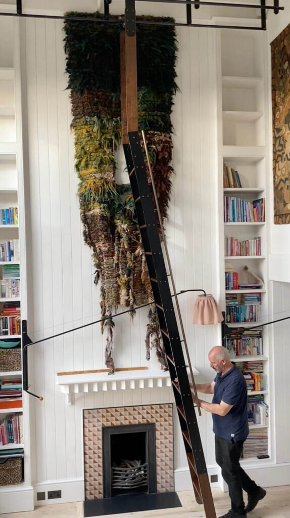 A man on a library ladder reaches toward a bookshelf in a room with white paneled walls. There is a large, textured tapestry hanging above a fireplace. Shelves on either side hold colorful books.