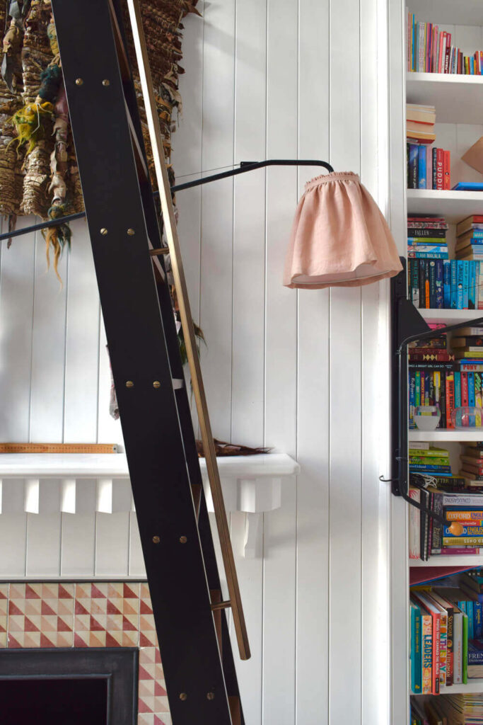 A cozy reading nook with a tall ladder leaning against white paneled walls, leading to shelves filled with colorful books. A soft pink lamp adds warmth, and a decorative piece hangs by the fireplace with a geometric pattern below.