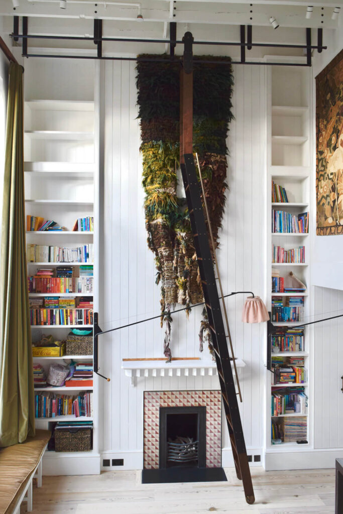 A cozy library features a white fireplace with bookshelves on either side, filled with colorful books. A tall ladder leans against a shelf. Above the fireplace hangs a large, textured tapestry in earthy tones.