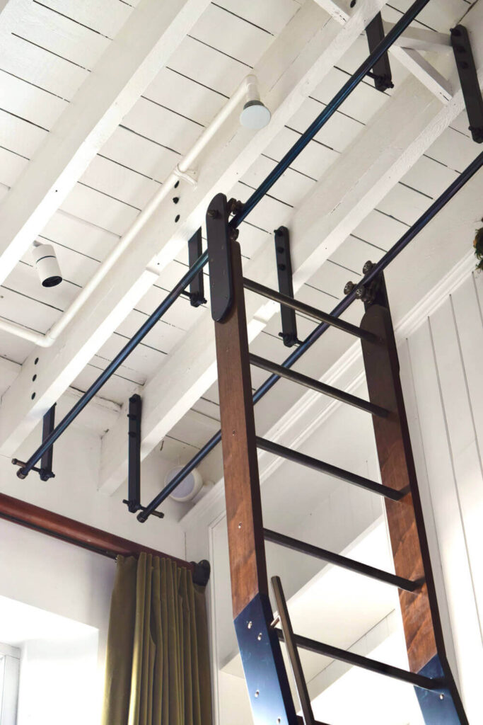 A wooden ladder with metal supports leads up to a white, open-beam ceiling in a well-lit interior. The ceiling features track lighting, and a green curtain is partially visible to the left.