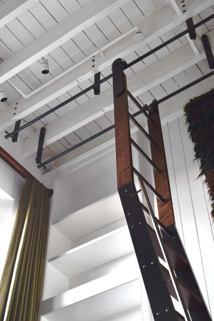 Wooden ladder leading up to a loft area against a white-paneled wall. The ceiling has exposed white beams. Shelves are visible on the left, and there are gold curtains at the edge of the image.