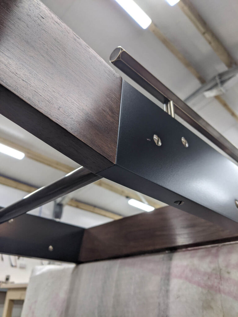 Close-up of a metal and wood structure, possibly part of a furniture piece or architectural element, with visible screws and beams. The background shows a workshop setting with bright overhead lights and exposed ceiling beams.