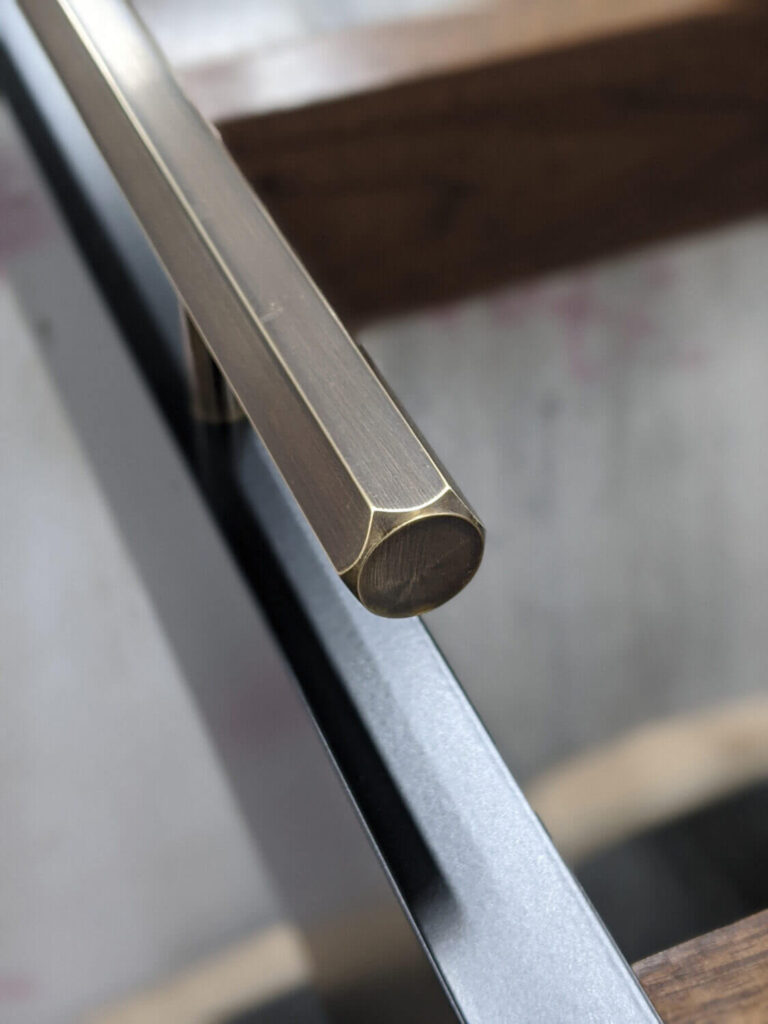 Close-up of a sleek, metallic handle attached to a glass surface. The handle has a brushed, bronze-like finish with a hexagonal shape, resting atop the edge of a transparent glass pane, possibly part of a door or table.