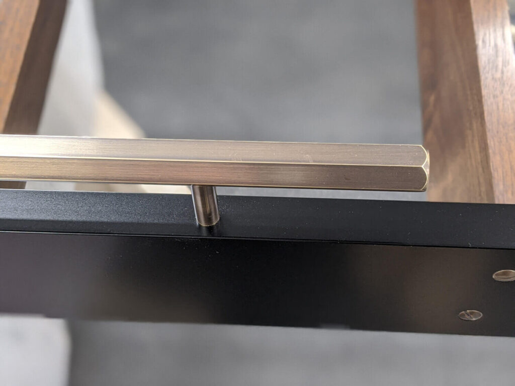 Close-up of a sleek, modern metal drawer handle mounted on a black finish panel with visible wood edges in the background. The handle is cylindrical with a brushed texture, attached by a metal rod. The surface has a minimalist design.