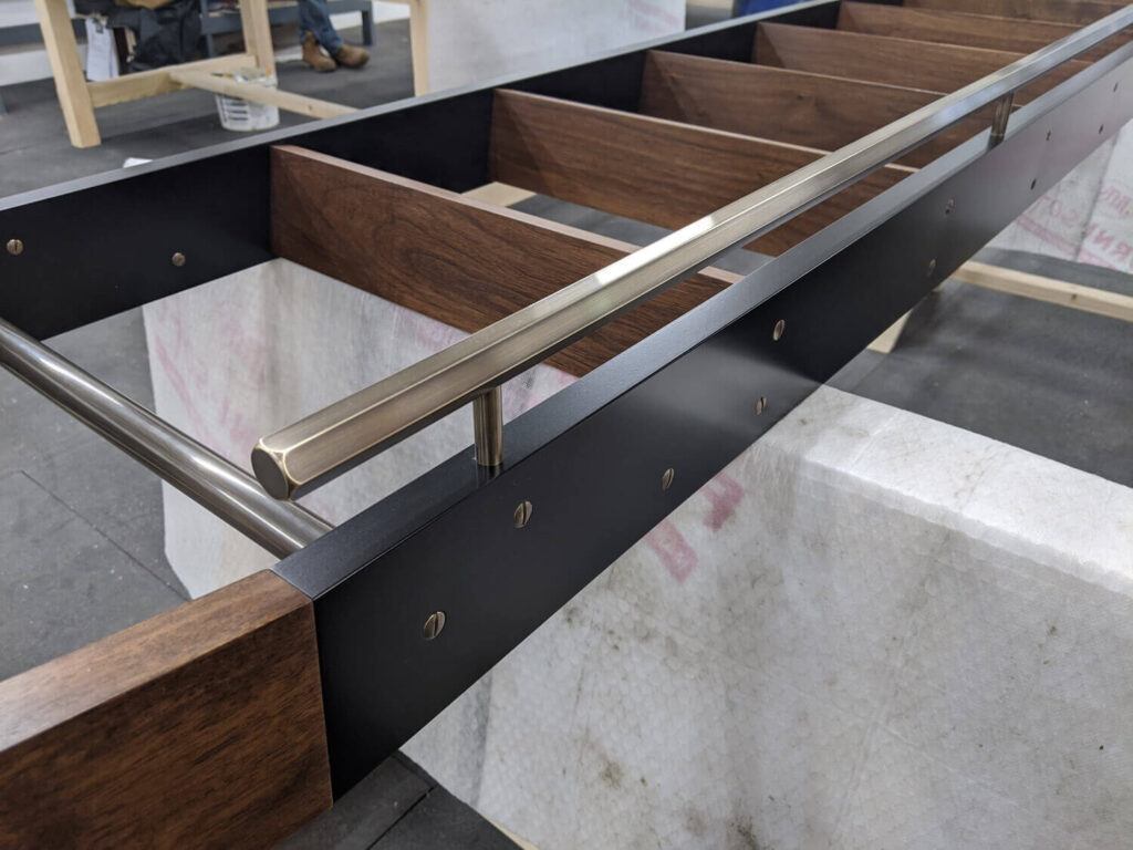 A close-up of a modern staircase under construction. The design features dark wood and black metal side panels with a sleek metal handrail. The steps are unfinished, and protective materials are visible underneath.