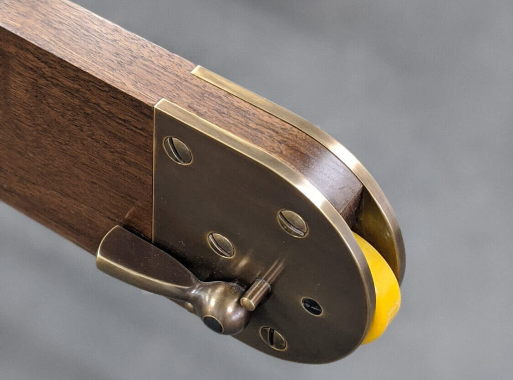 Close-up of a wooden library ladder castor with a brass fitting and a yellow rubber wheel. The fitting is secured with several screws and features a small brass lever. The background is slightly blurred, focusing on the hardware detail.