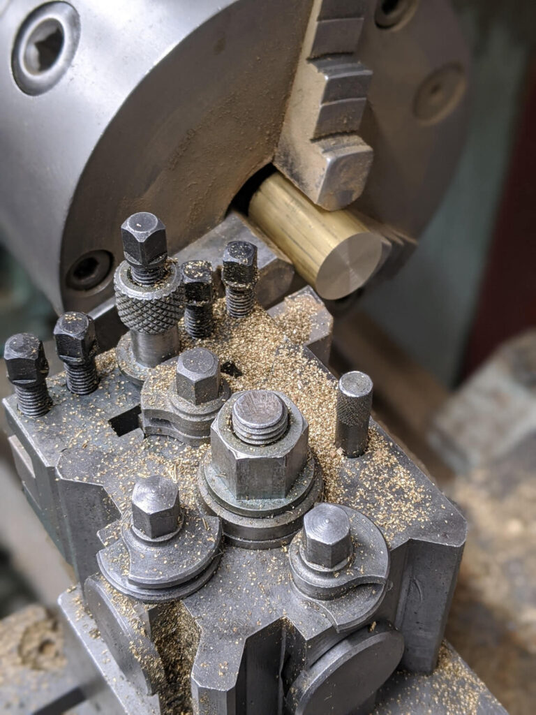 Close-up of a metal lathe machining a brass rod. The rod is clamped in the chuck, and metal shavings are scattered on the lathes tool post, highlighting the precision and mechanics of the machining process.