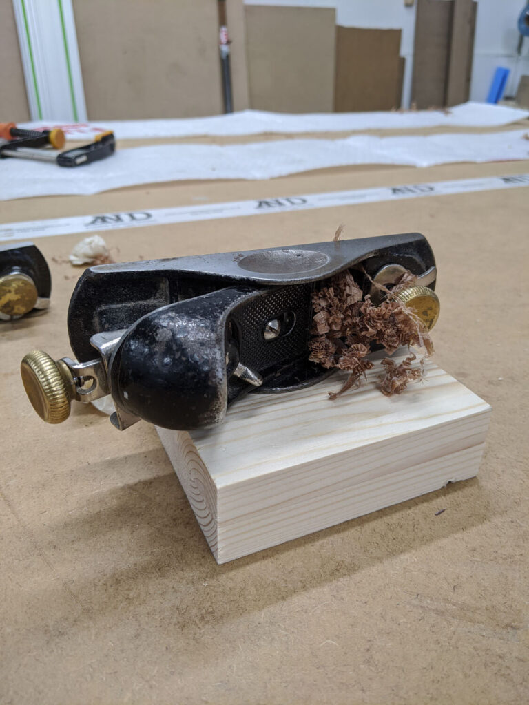 A hand planer rests on a piece of wood, with wood shavings piled around the blade. In the background, other woodworking tools and supplies are visible on a workbench.