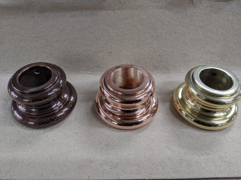 Three metallic curtain rod finials are displayed on a beige surface. From left to right, they are in bronze, copper, and brass finishes, featuring a rounded, ribbed design.