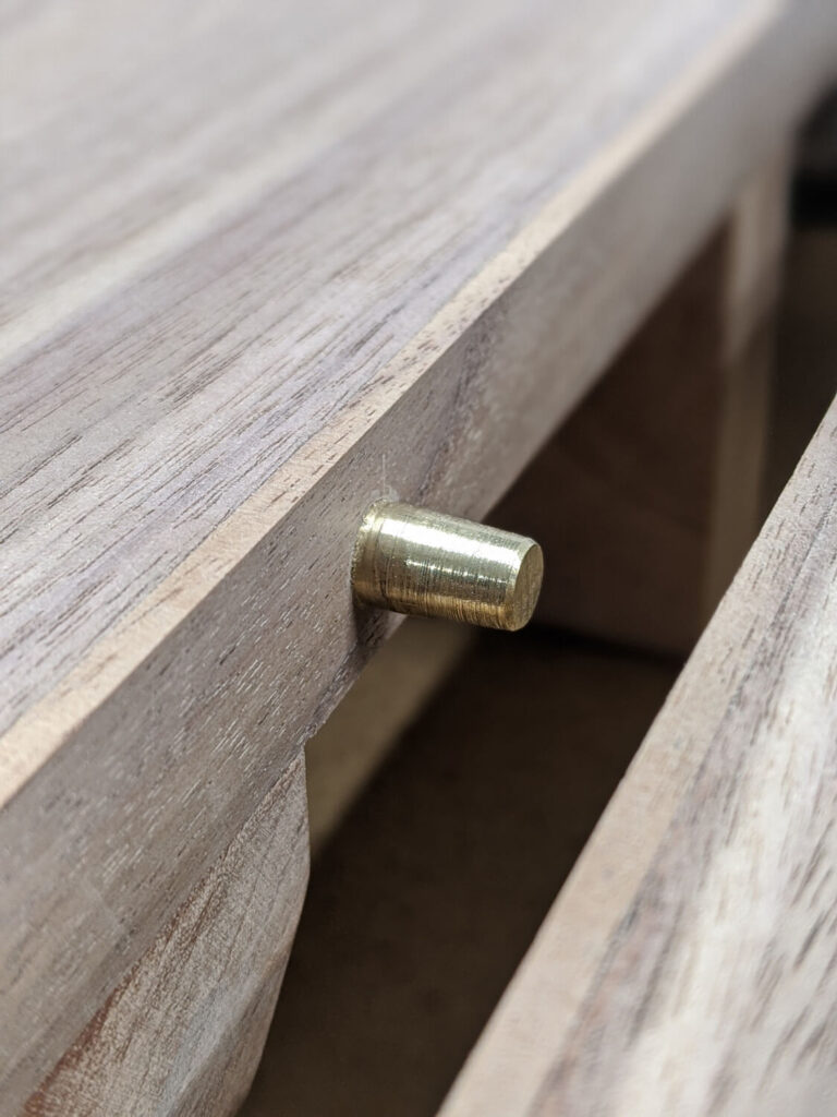 Close-up of a wooden frame with a cylindrical brass peg partially inserted into a horizontal groove.