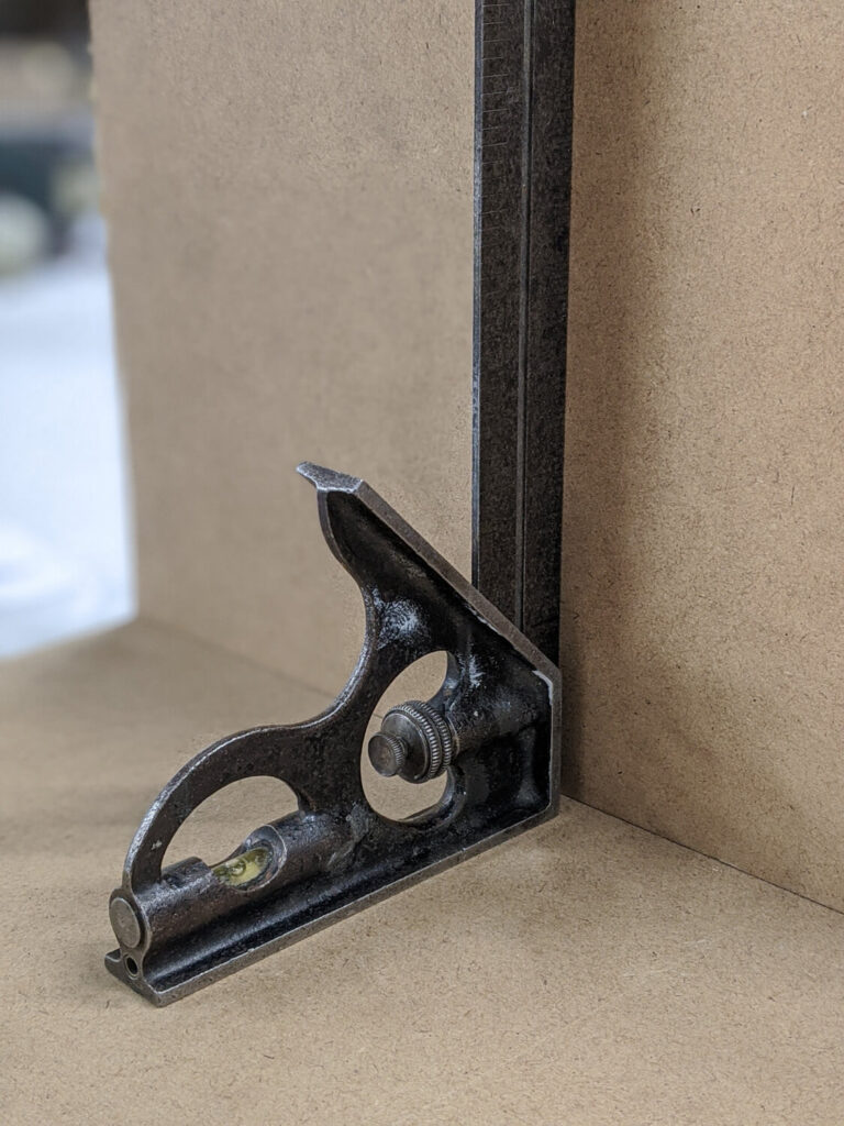 A close-up of a metal combination square placed against a wooden board, used for measuring and marking angles in carpentry or metalworking. The tool includes a spirit level and a ruled edge for precision.