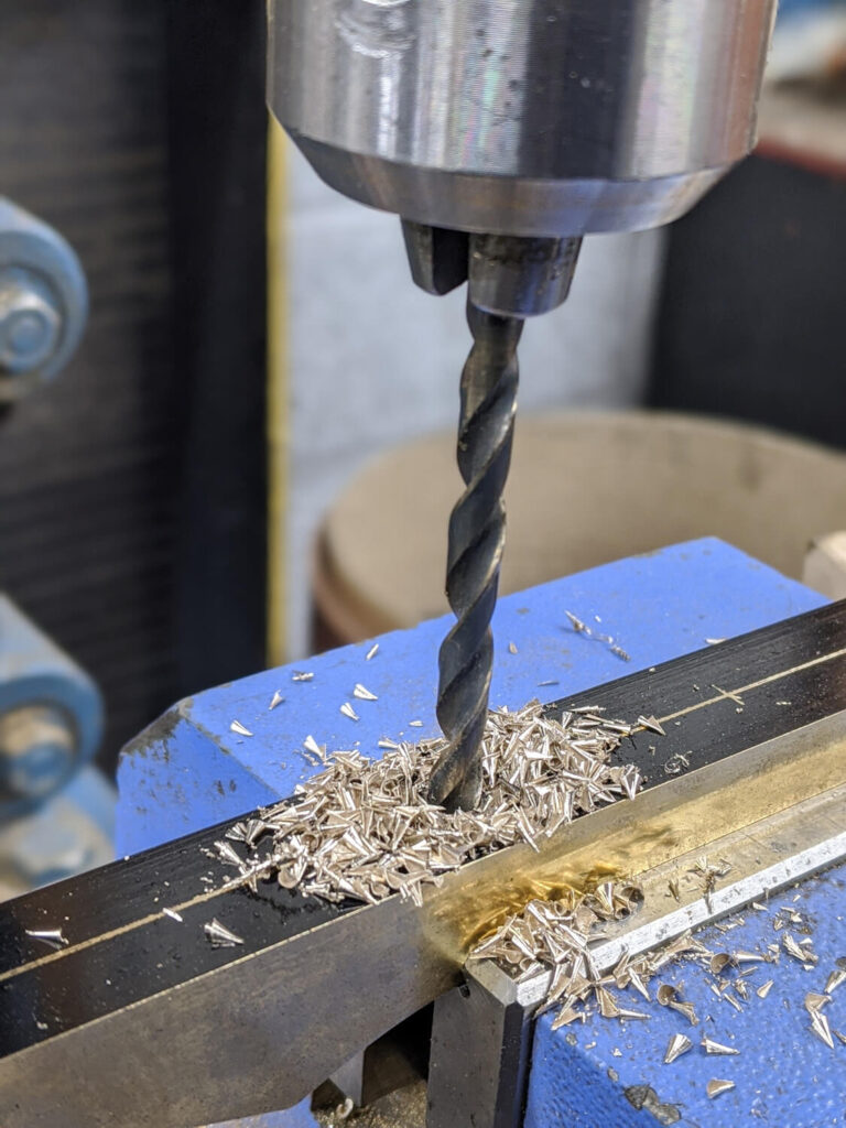 A close-up of a drill press in action, drilling into a metal surface. Metal shavings surround the drill bit as it cuts through the material securely held in a blue vise. The background is blurred, emphasizing the machining process.