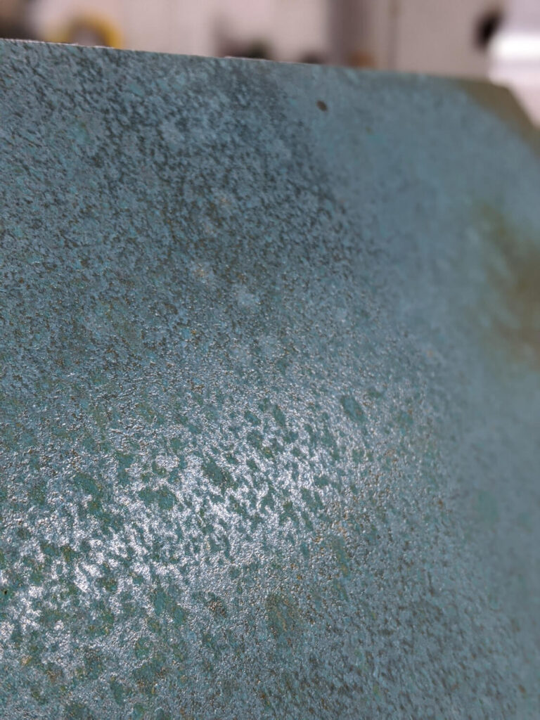 Close-up of a textured, greenish-blue metallic surface with patches of rust and wear, capturing the light and showing variations in color and texture.