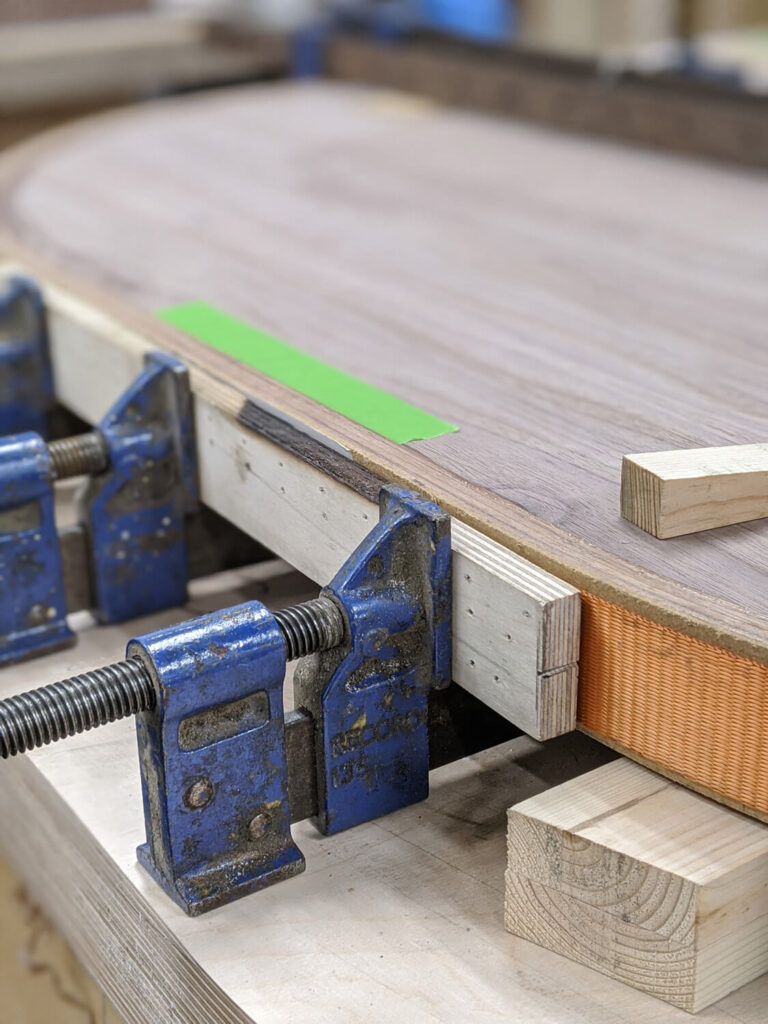 A woodwork project in progress shows an oval wooden tabletop secured with large blue clamps on a workbench. The edge is reinforced with wood pieces and a green tape is visible on the surface.