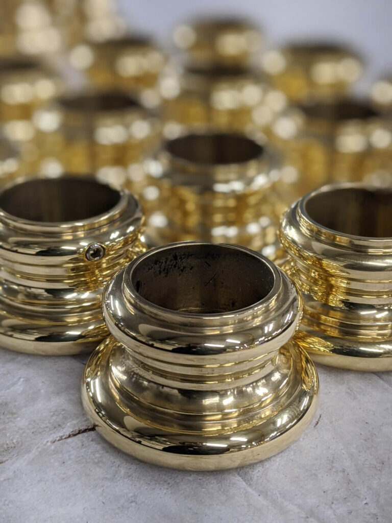 Close-up of several polished brass metal rings, with shiny, reflective surfaces. They are arranged on a textured, light-colored surface, creating a pattern of gleaming, concentric circles.